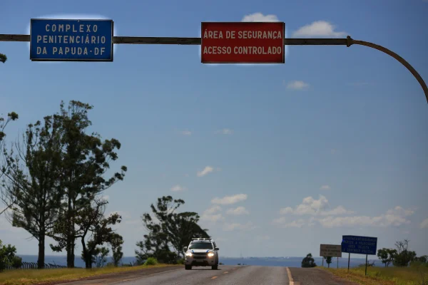 Imagem colorida de duas placas coladas a um poste. As placas contêm os escritos: 'COMPLEXO PENITENCIÁRIO DA PAPUDA-DF' e 'ÁREA DE SEGURANÇA ACESSO CONTROLADO'