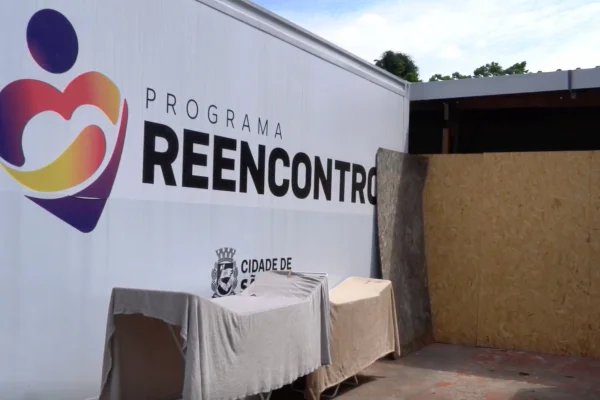 Imagem colorida mostra parede branca com logotipo do Programa Reencontro em um pátio com panos estendidos e uma parede de madeirite