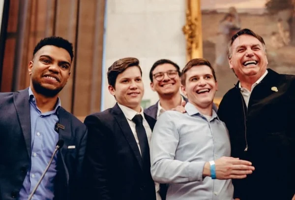 Imagem colorida mostra Fernando Holiday, homem negro de terno cinza e camisa azul, um grupo de jovens e o ex-presidente Bolsonaro, homem branco de jaqueta preta. Todos sorriem na imagem - Metrópoles