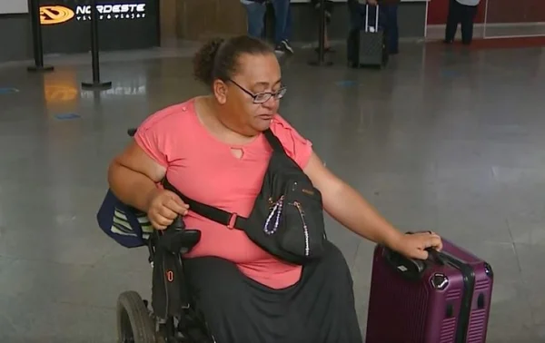foto colorida de mulher cadeirante de blusa rosa segurando mala em rodoviária