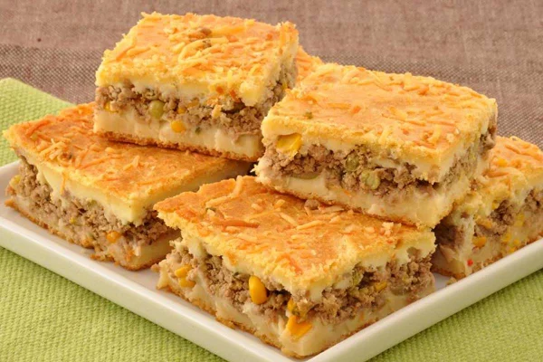 A foto mostra seis pedaços quadrados de torta de carne moída em uma prato branco e uma mesa verde clara
