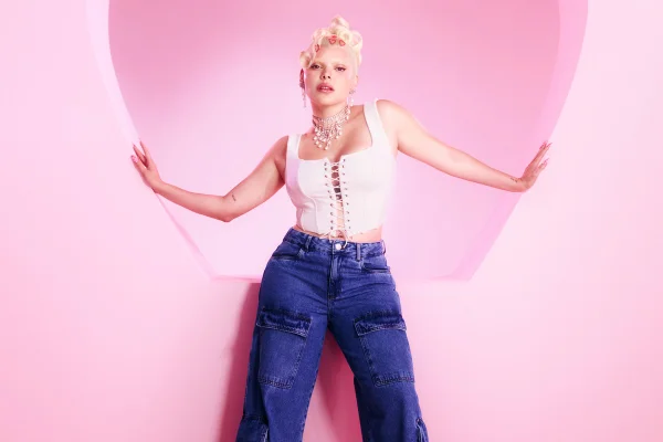 Campanha de divulgação da coleção de roupas da C&A em parceria com a cantora Duda Beat. Na foto, é possíverl ver Duda Beat, uma mulher branca, jovem e com cabelo loiro platinado em um fundo rosa. Ela veste um top corset branco e uma calça jeans