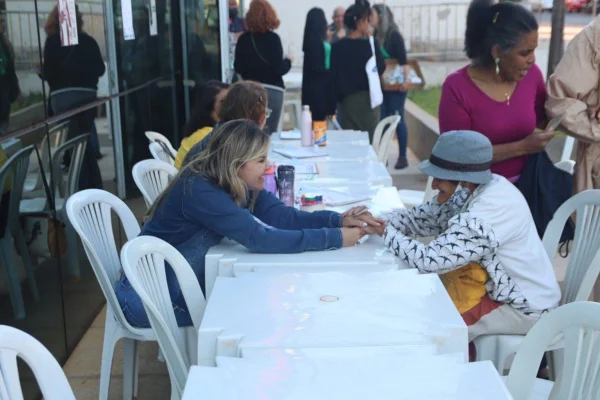 Imagem colorida de duas mulheres sentadas frente a frente, separadas por uma mesa branca