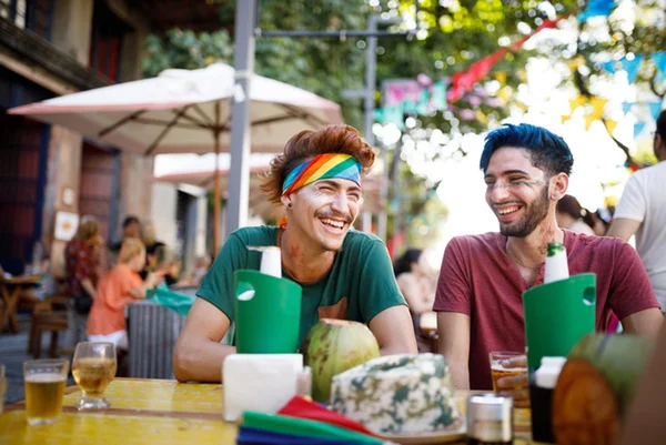 Dois homens com maquiagem de Carnaval sentados em mesa de bar tomando cerveja e água e coco - Metrópoles