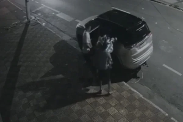 Imagem em preto e branco de dois homens com sacolas de lixo ao lado de carro de luxo. Metrópoles