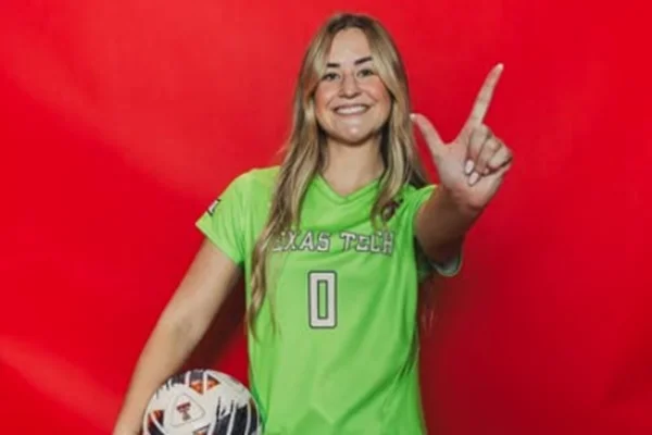 Foto colorida de jogadora de futebol, segurando a bola, fazendo síbolo do time, em frente a uma parede vermelha - Metrópoles.