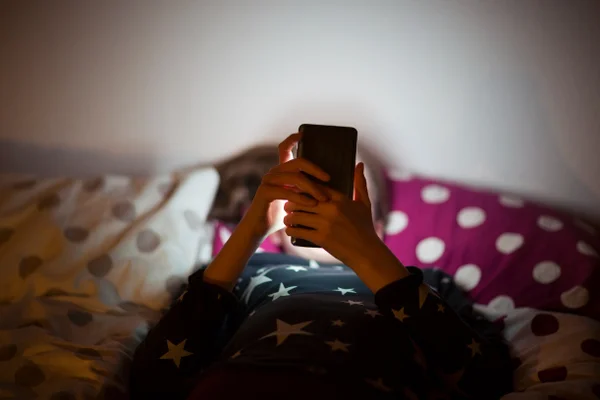 Imagem colorida de criança deitada em uma cama mexdendo no celular - Metrópoles