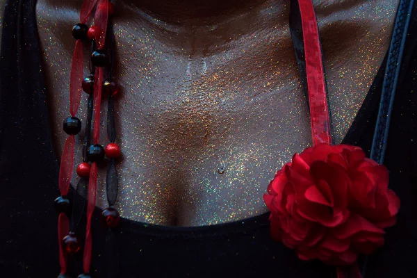 Foto colorida em close do colo de uma mulher suada com glitter na pele, uma blusa preta e uma rosa vermelha pendurada - Metrópoles