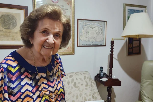 imagem colorida mostra senhora de 90 anos. ela é branca, tem os cabelos claros, e sorri para a foto. usa um vestido colorido e está em uma sala