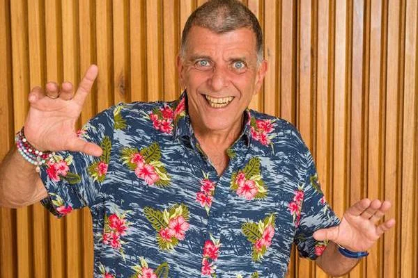Jorge Fernando em frente a uma parede de madeira, usando camisa florida - Metrópoles