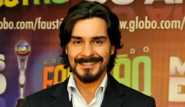 Na imagem colorida, um homem está posicionado no centro. Ele usa casaco preto, blusa branca, tem cabelos escuros, bigode e sorri para a camera