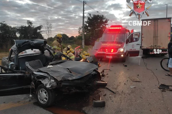 Imagem colorida mostra carro preto destruído após acidente. Ao lado dele, um grupo de bombeiros com uniformes amarelos trabalham no local e, ao fundo, há uma ambulância e um pequeno caminhão – ambos parados