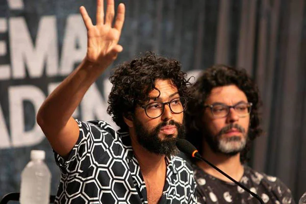 homem de barba e oculos fala em microfone ao lado de outro homem