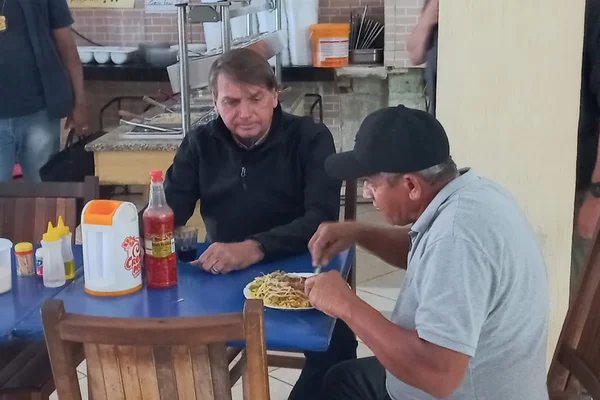 Bolsonaro observa caminhoneiro comer em restaurante no Distrito Federal. Ele usa jaqueta preta e o homem, camiseta branca e boné - Metrópoles