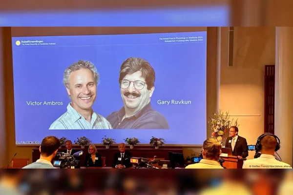 Imagem mostra um auditório e no telão as fotos de Victor Ambros e Gary Ruvkun que ganharam o prêmio nobel de 2024. Metrópoles