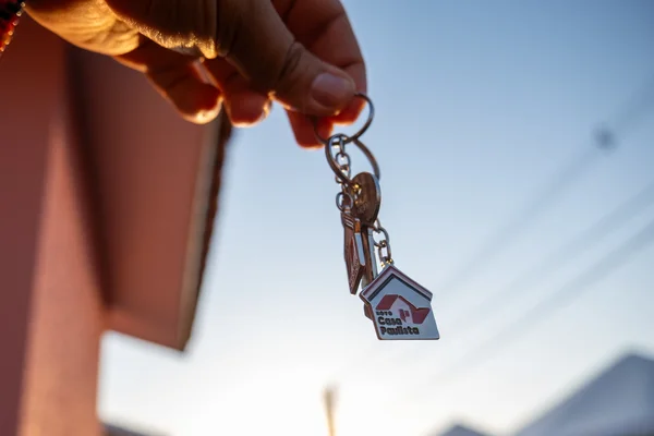Imagem em close mostra uma mão segurando um chaveiro com o formato de uma casa. Ao fundo, desfocado, aparece um ambiente externo com luz natural, sugerindo o momento simbólico da conquista de um novo lar