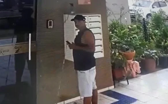 Homem de boné, regata preta e bermuda branca em frente a entrada do prédio