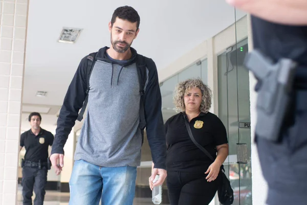 Homem de casaco cinza e mochila caminha ao lado de policial civil