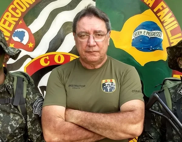 Homem com camiseta marrom, cabelos grisalhos e óculos, de braços cruzados - Metrópoles