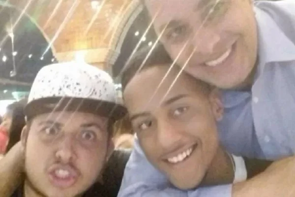 Foto com três homens. Um de boné e camisa preta, um sorrindo com cabelo preto e baixo e outro com camisa social azul e sorrindo. - Metrópoles