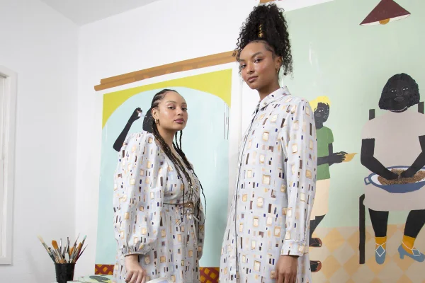 Duas mulheres jovem e negras, uma com cabelo cacheado e outra com tranças, posando para foto com um vestido e um conjunto de saia e blusa. As roupas são azuis claras, possuem pequenos desenhos colorudos e são fruto de uma coleção especial entre a C&A e a artista plástica Larissa de Souza. No fundo é possível ver uma obra da artista. - Metrópoles