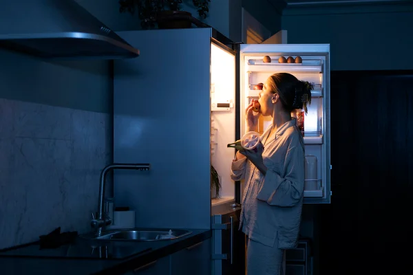 Imagem mostra mulher comendo enquanto olha dentro da geladeira - Metrópoles