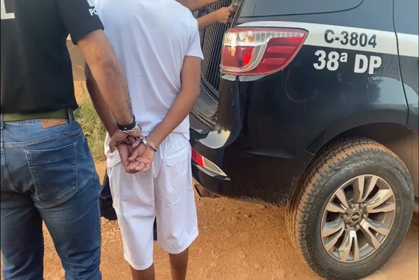 Dois homens de costas, um dele, com camisa preta da PCDF, segura o segundo homem, vestido de branco e algemado, para colocá-lo em carro preto da 38ª DP - Metrópoles
