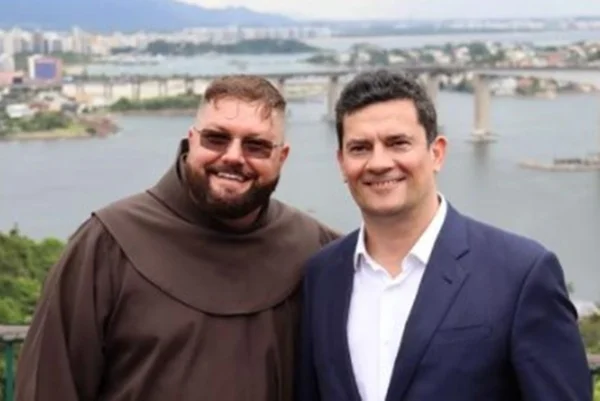 Homem com roupa de frei e óculos escuro ao lado de homem de terno e paisagem ao fundo