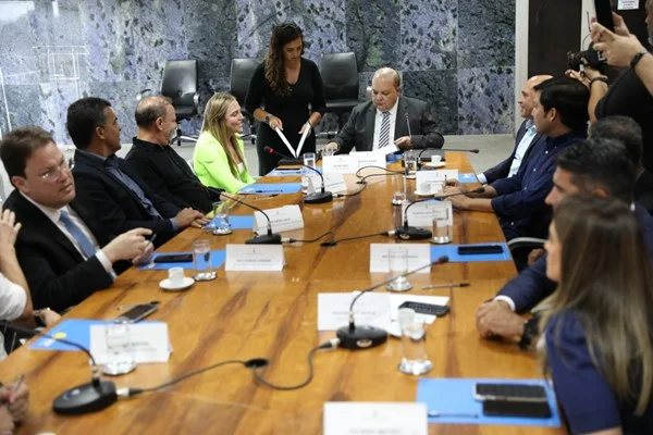 Fotografia colorida de mesa de reunião com pessoas de terno e mulher de blazer verde