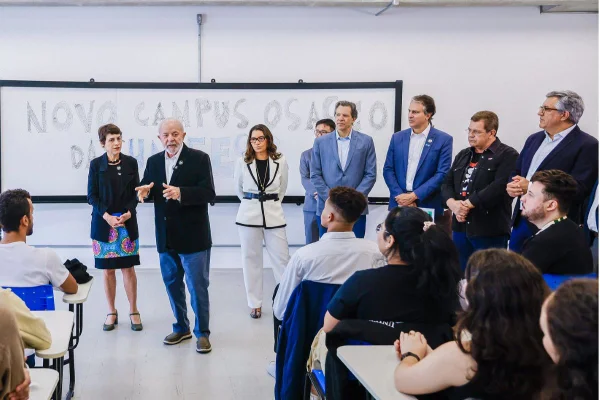 Homem branco de cabelos brancos vestindo blazer preto está em frente a quadro de sala de aula; há pessoas em pé ao redor e outras sentadas em carteiras - Metrópoles