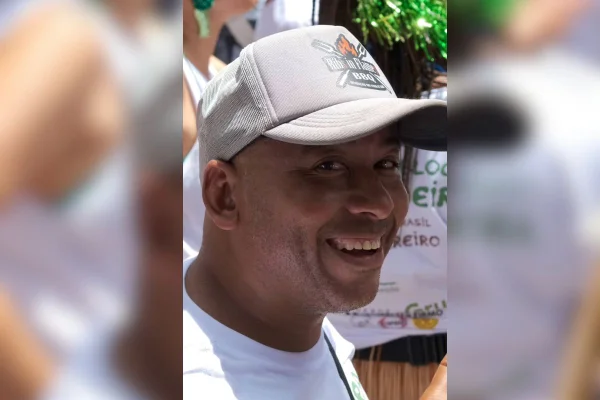 imagem colorida mostra homem negro sorrindo. ele usa um boné de cor cinza e uma camiseta branca