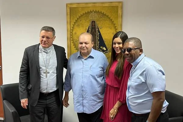 Fotografia colorida de três homens e mulher sorrindo para foto com imagem santa ao fundo