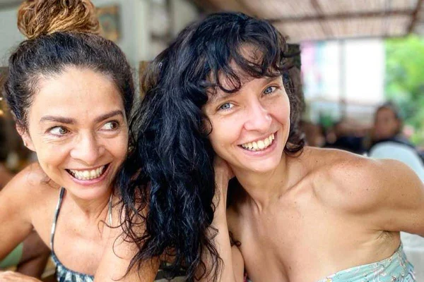 mulher braca sentada com cabelo presso em coque sorri e olha para o além enquanto a outra posa ao lado de cabelos soltos sorrindo - metrópoles