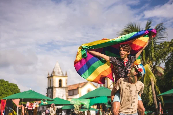 Um homem levanta uma bandeira LGBTQIAPN+ enquanto outro o segura nas costas, sorrindo. Ao fundo, aparece uma feira ao ar livre e prédios antigos - Metrópoles