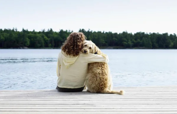 Foto colorida de mulher sentada na beira do pier abraçada com o cachorro - Metrópoles