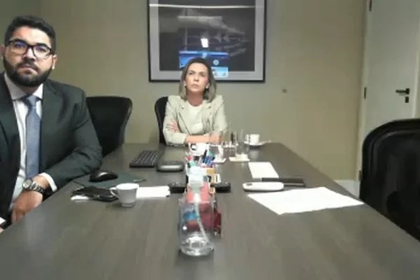 Jornalista acusada de chamar colega negra de "carvão" durante audiência ao lado de seu advogado. Ambos aparecem sentados em mesa, olhando para frente - Metrópoles