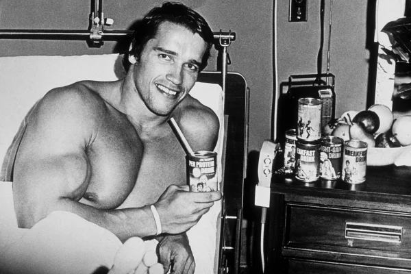 Imagem em preto e branco mostra o ator Arnold Schwarzenegger jovem tomando um energético de café da manhã - Metrópoles