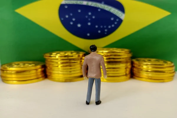 Imagem da bandeira do Brasil com várias moedas e um bonequinho olhando para elas - Metrópoles