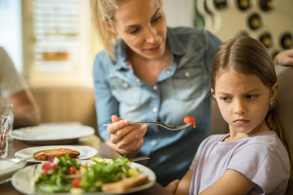Imagem de criança sentada à mesa, se recusando a comer tomate oferecido por mulher - Metrópoles
