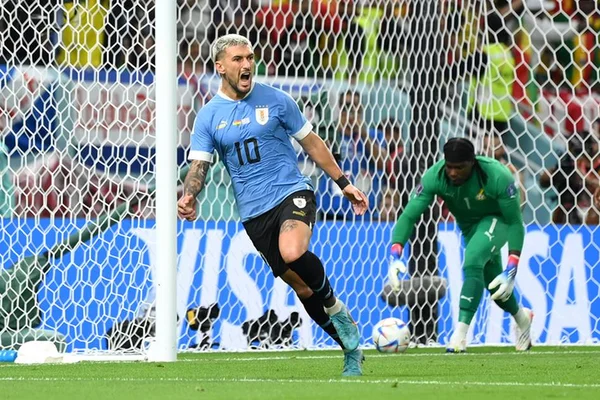 Jogador Arrascaeta, um homem branco e de cabelo descolorido correndo para comemorar após marcar um gol. Vestindo roupa azul do time do Uruguai, ele está gritando. Ao fundo, aparece um goleiro de roupas verdes tentando pegar uma bola que está dentro do gol