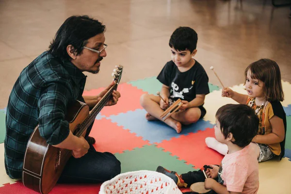 Homem com violão sentado em tapete de eva com três crianças segurando instrumentos - metrópoles