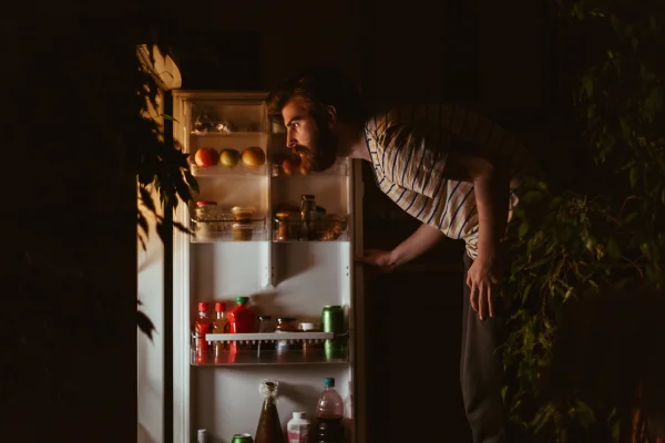 Fotografia mostra homem no escuro olhando dentro de geladeira iluminada - Metrópoles