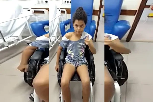 Criança de roupa azul sentada em cadeira de rodas e fazendo sinal de "tudo bem" com a mão