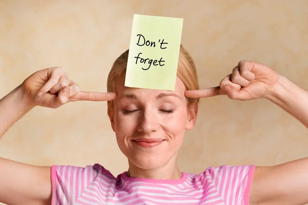 Mulher loira jovem com os dedos apontando para a cabeça com um post it na testa com a frase don't forget