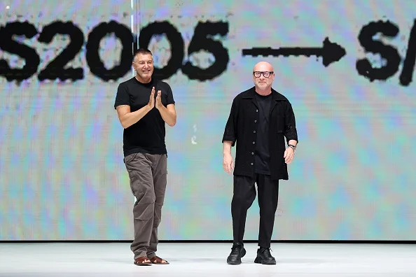 Os estilistas Domenico Dolce e Steffano Gabbana em passarela durante o Milan Fashion Week. Eles sorriem para a plateia diante de um telão - Metrópoles