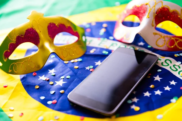 Na foto, celular sobre bandeira do brasil junto de máscaras de carnaval