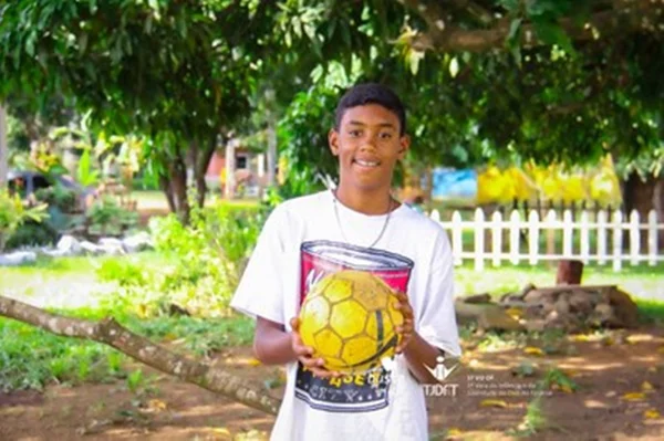 Menino negro com camisa branca segura bola amarela em área com árvores