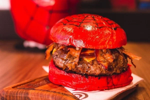 Na fotografia vemos um hambúrguer com pão vermelho no qual está desenhado uma teia de aranha. No meio, dá para ver a carne, pedaços de bacon e cebola caramelizada