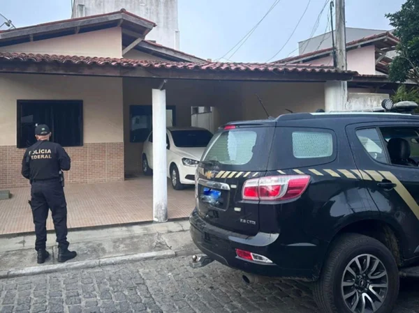 Imagem colorida de agente e viatura da PF diante de uma casa com um carro branco na garagem - Metrópoles