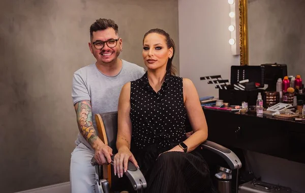 Paulo Lobo está com camisa cinza clara e sentado atrás da modelo Bia Fernandes. Ambos olham para a câmara para mostrar o resultado da maquiagem rocker
